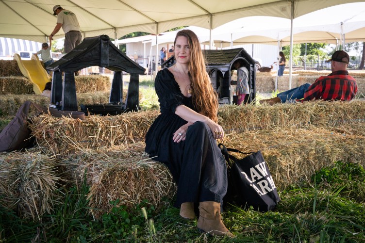 A woman in a black dress sits on a hay bale next to a Raw Farm tote bag under a white tent, with other people in the background.