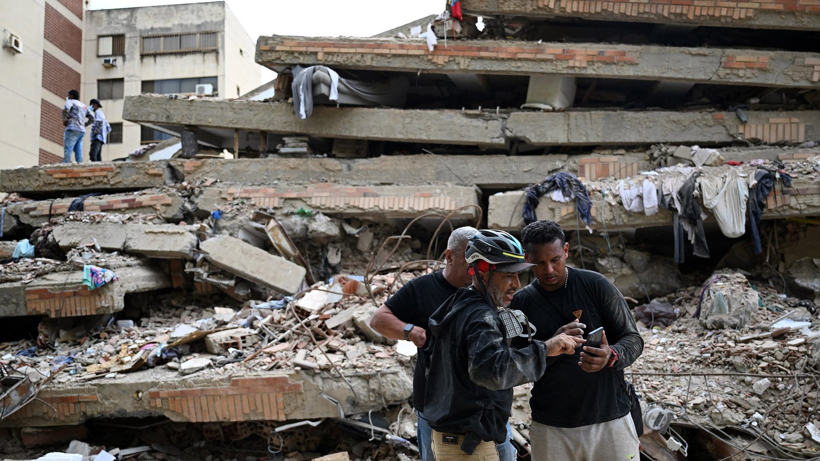 Two men examine a phone amidst the rubble of a collapsed building, with debris and scattered clothing around them