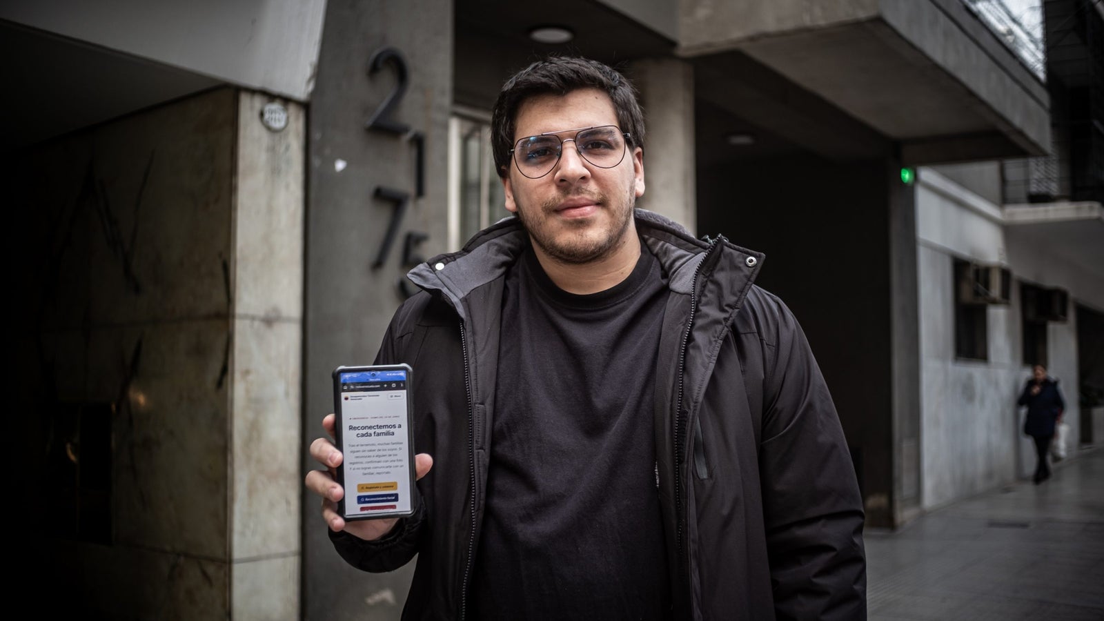 A man in a black jacket stands outside a building, holding a smartphone displaying a website. The building has the number 217 on it