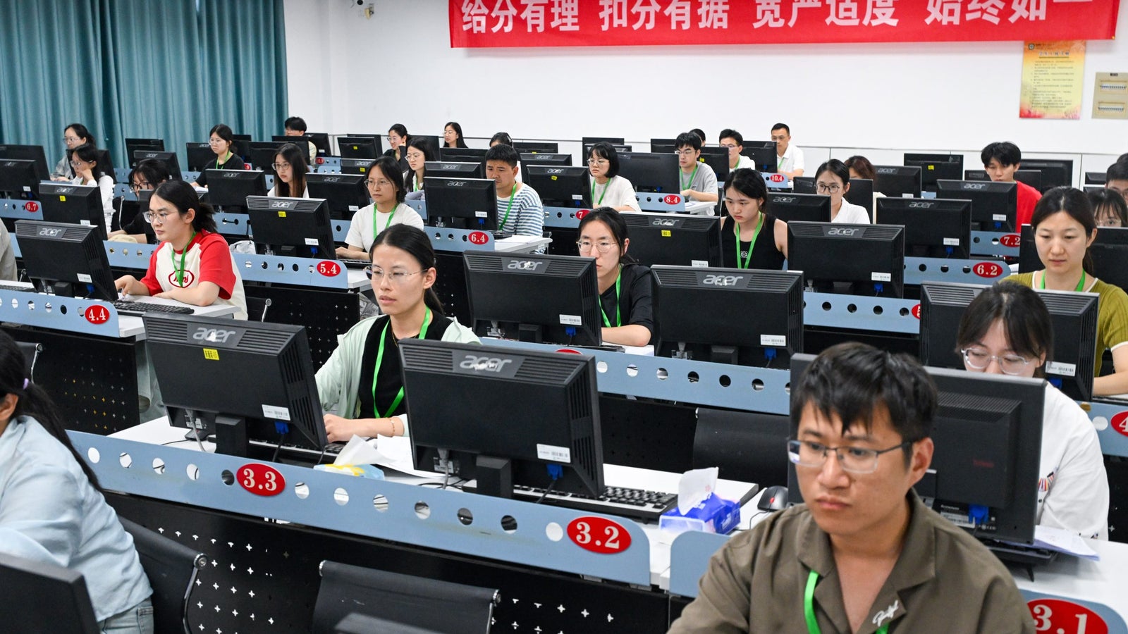 A classroom filled with students focused on computers, each seated at desks with monitors and identification tags