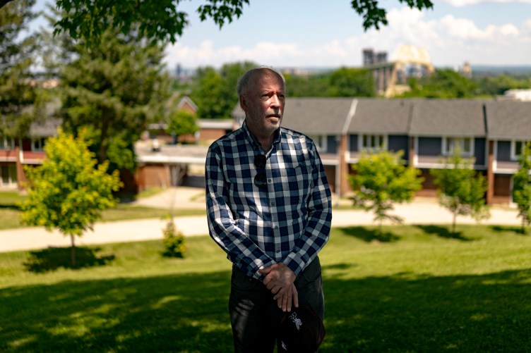 An older man with graying hair and a beard stands outdoors in a grassy area, wearing a blue-and-white plaid button-up shirt and dark pants. He rests his hands together in front of him, holding a dark cap.