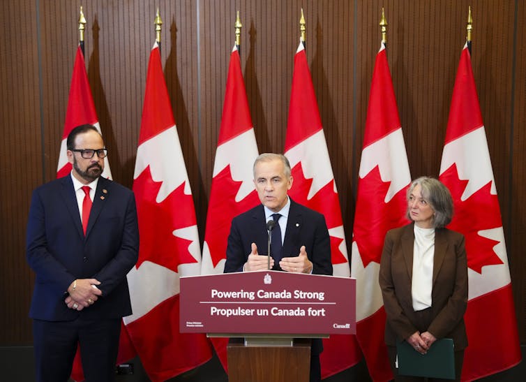 A man in a suit at a podium labelled Powering Canada Strong, flanked by a man and a woman in front of a row of Canadian flags.