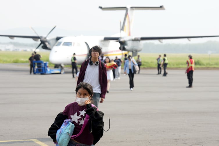 a girl wearing a face mask and other people walk from a plane on tarmac
