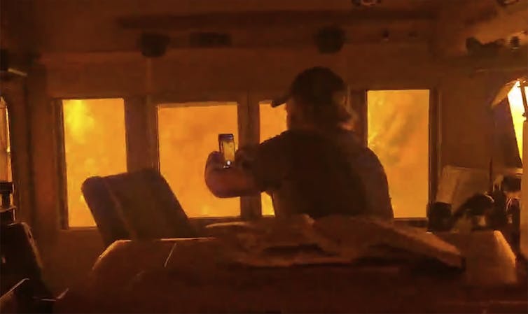 A man in the cab of a train surrounded by flames as he holds up a phone.