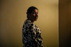 A woman with dark hair stands in a darkened hotel corridor.