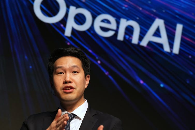 A man in a suit speaks passionately with an OpenAI banner behind him