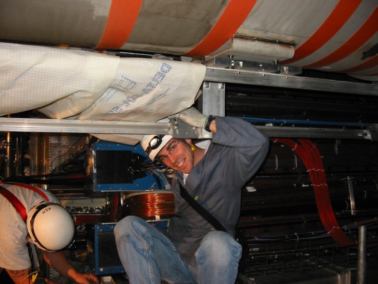 A young man wearing a helmet and a headlamp is wedged between a metal structure covered in wires.
