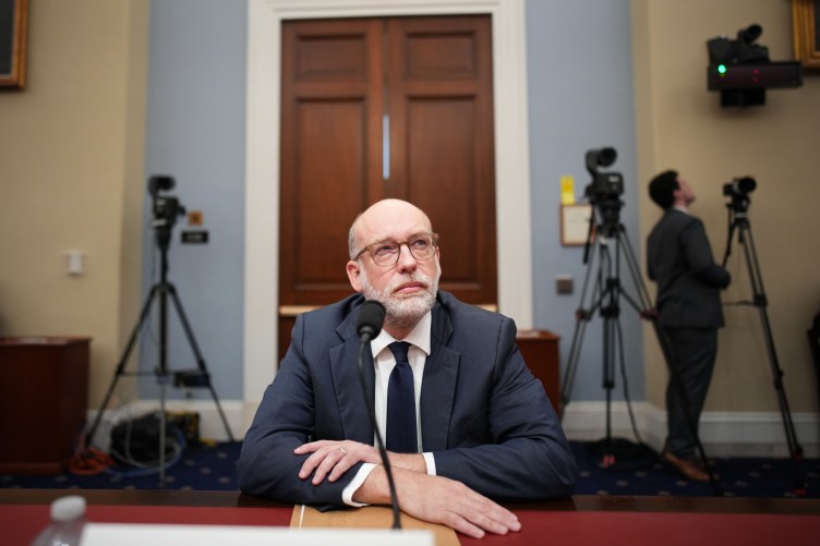 A man in a suit and glasses with his arms crossed looks to his upper left. He is sitting at a desk with a microphone in front of him. Behind him are big doors, three tripods and a man setting up a camera.
