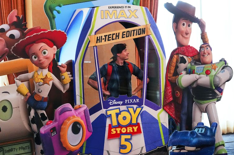 A person peeks their head through a life-sized movie display showing an astronaut and cowboy with the words 'Toy Story 5' and 'high-tech edition.'