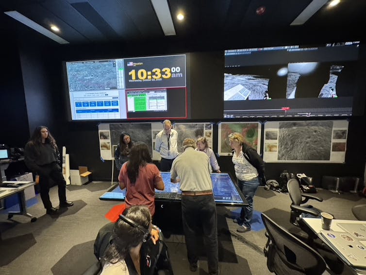 A group of people work together around a table in a room with large screens and equipment.