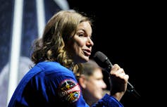 A woman with wavy dark blonde hair speaks into a microphone.