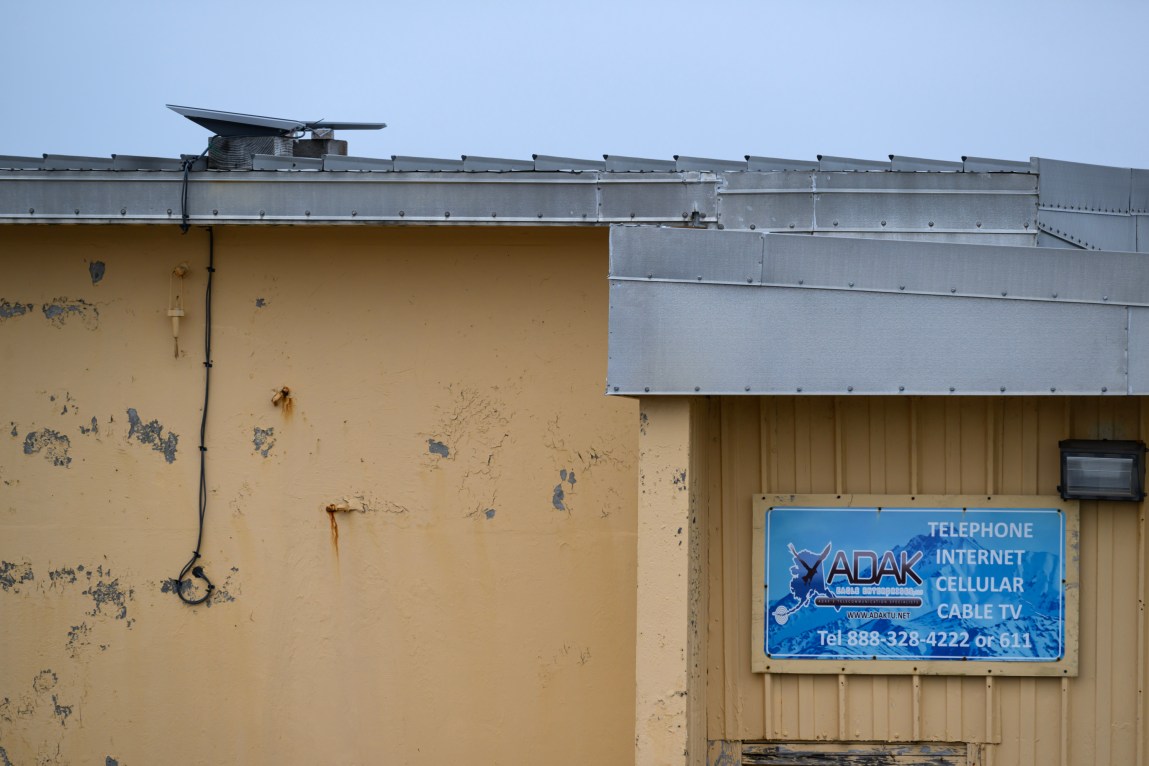 A building with yellow, peeling paint and a gray roof with a sign that says “Adak: Telephone, Internet, Cellular, Cable TV.”
