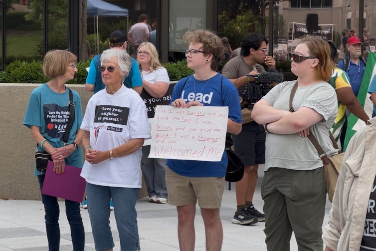Four women stand outside with signs and shirts that say things like, 'Love your neighbor as yourself.'