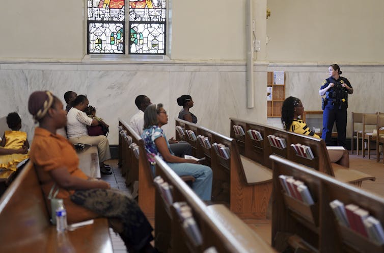 A woman in a blue uniform stands at the side of a sanctuary, in front of wooden pews with a few people seated in them.