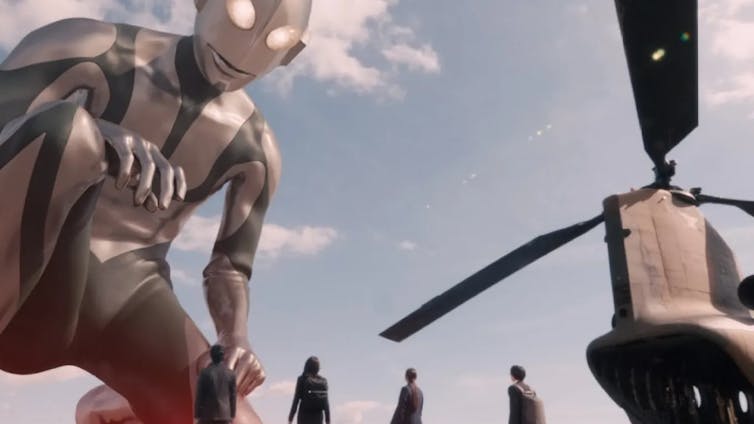 A giant chrome robot bends down on one knee to stare at four small human figures.