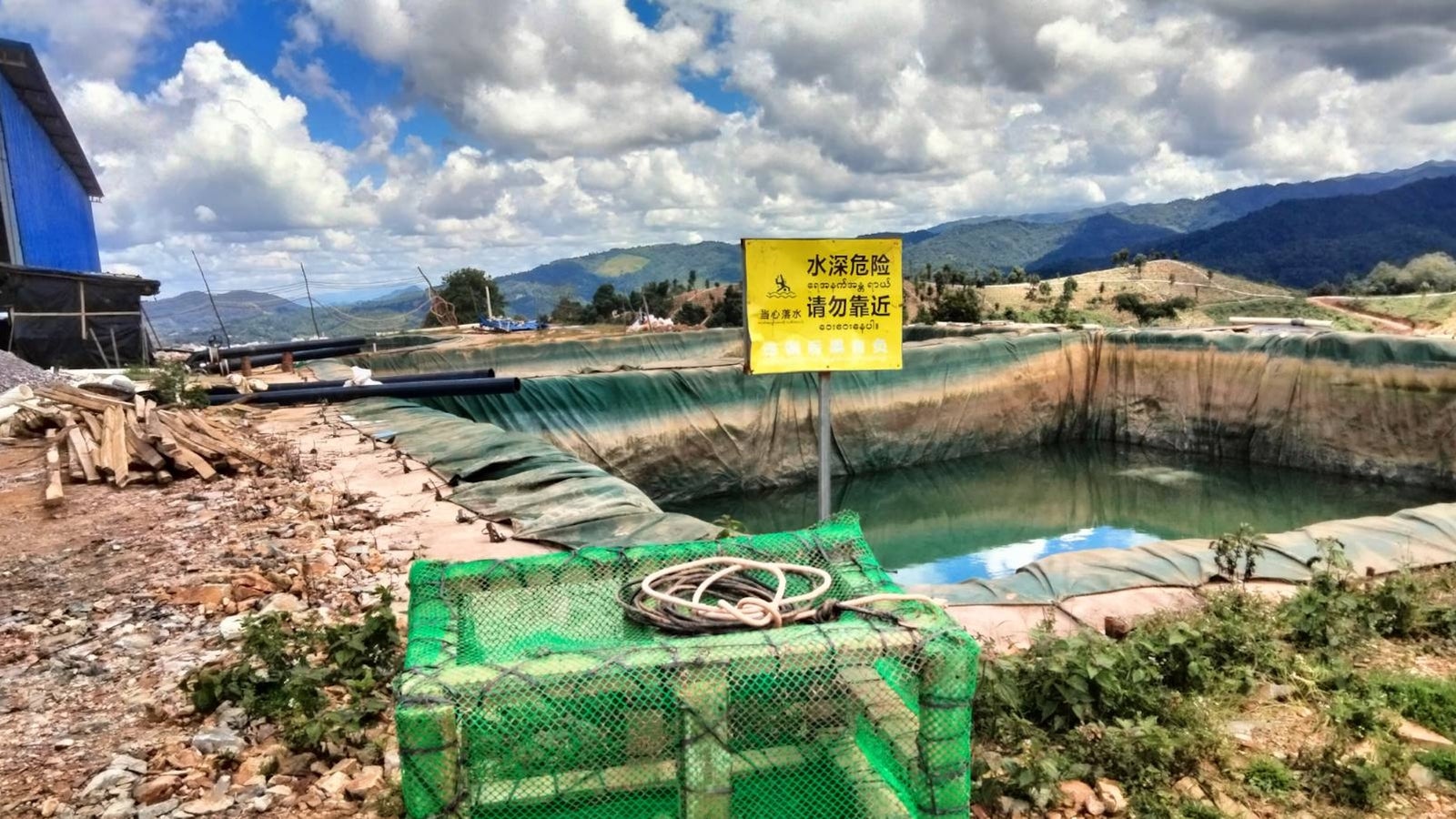 A green netted box with a coil of rope sits beside a large, fenced pond with a warning sign in a rural landscape