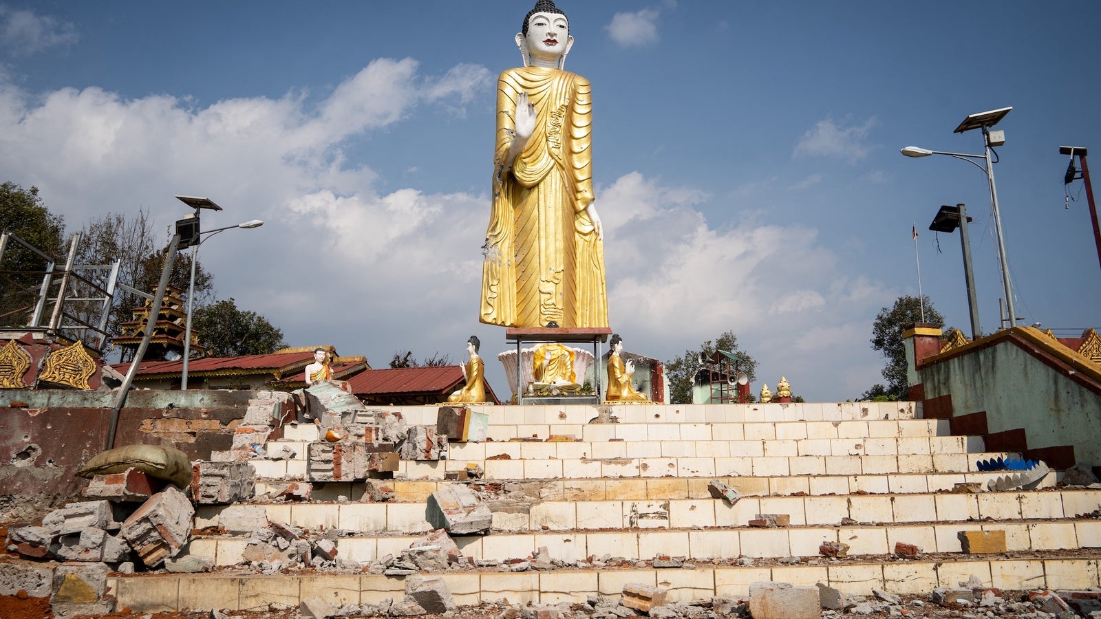A large golden Buddha statue stands on a staircase with debris and damaged structures around, under a partly cloudy sky