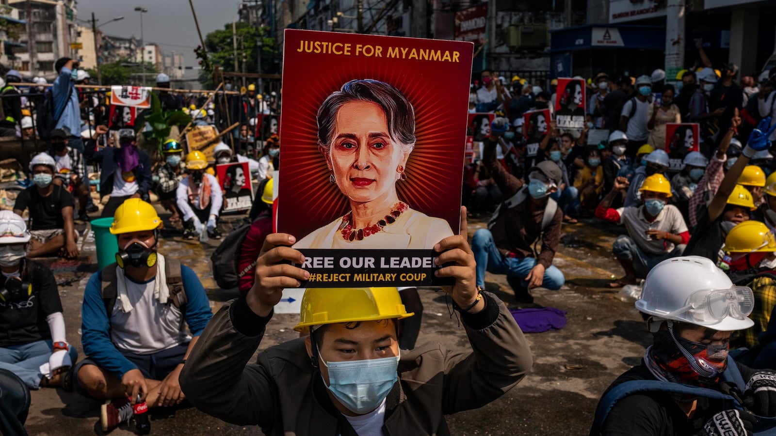 Protesters in hard hats demonstrate for justice in Myanmar, holding signs that demand the release of their leader