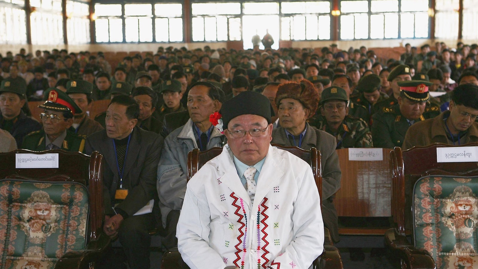 A crowd of seated individuals in a large hall, with a man in a white embroidered coat and glasses at the forefront