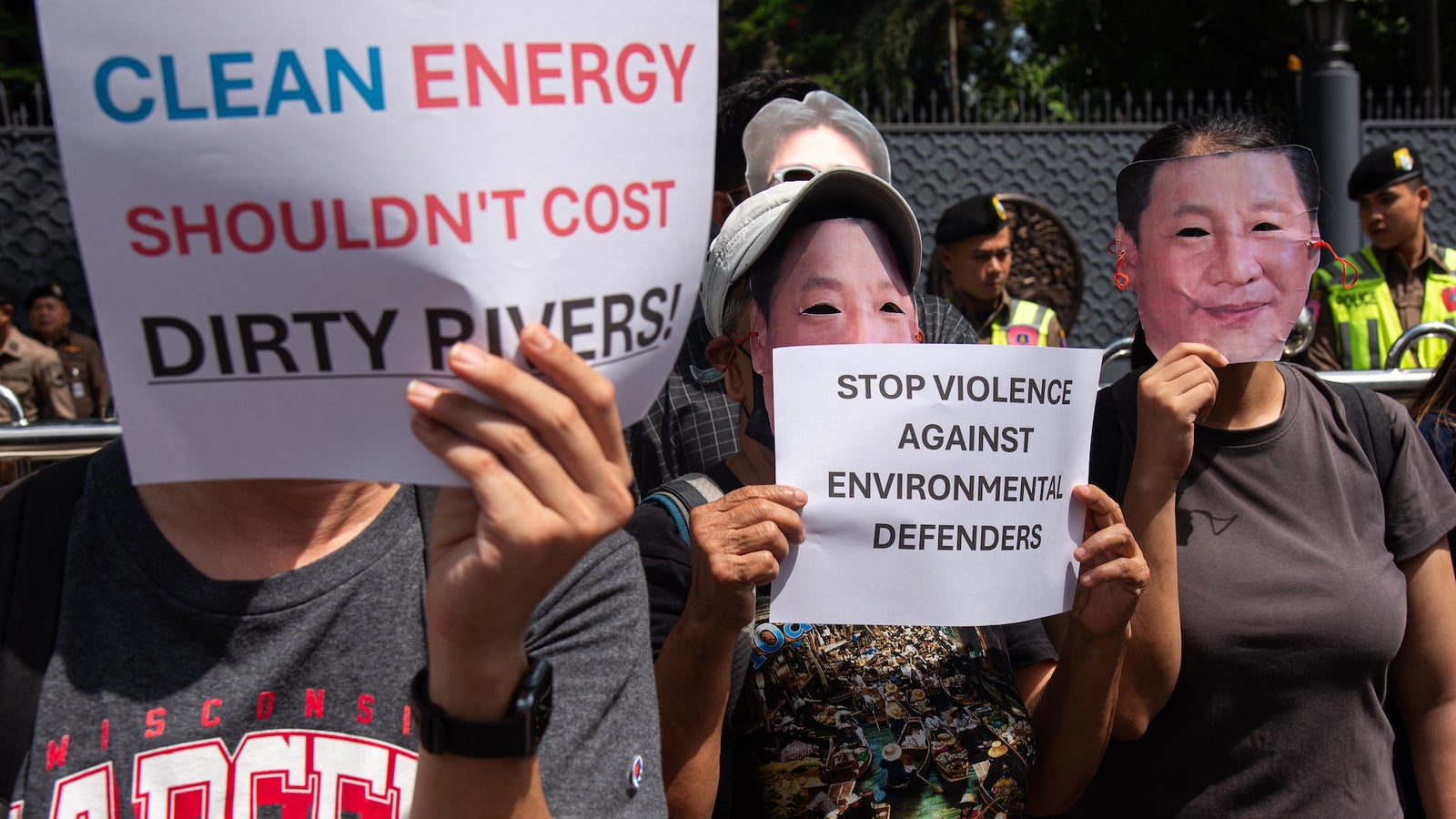 Protestors holding signs advocating for clean energy and against violence towards environmental defenders, wearing masks with faces