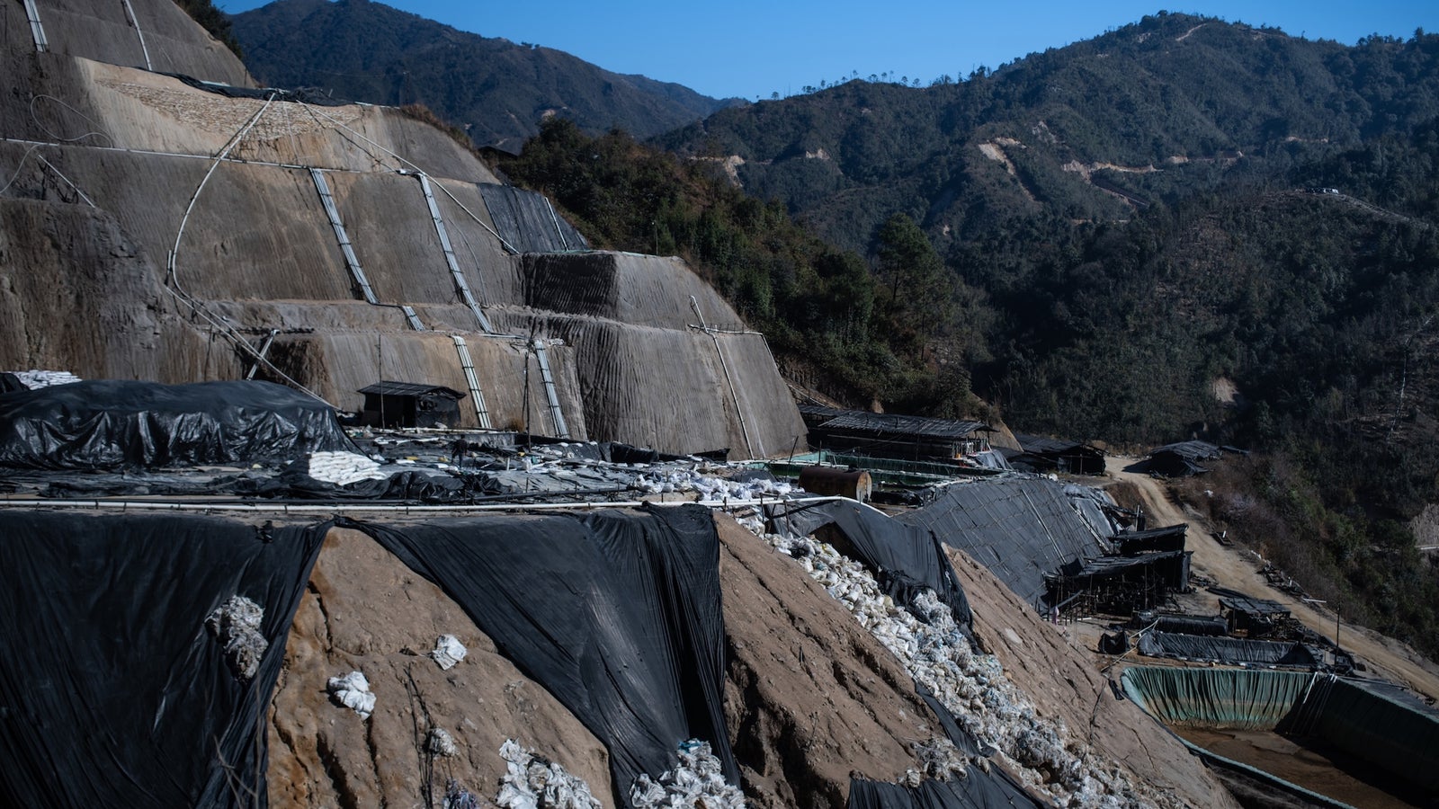 A landfill site with sloped layers covered in black tarps, surrounded by mountains under a clear blue sky