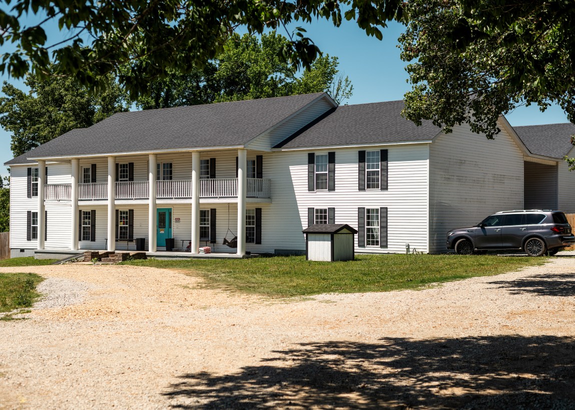 A white, two-story colonial-style building with multiple pillars and a balcony. A grey car is parked beside the building. Trees cast shadows on the dirt driveway.