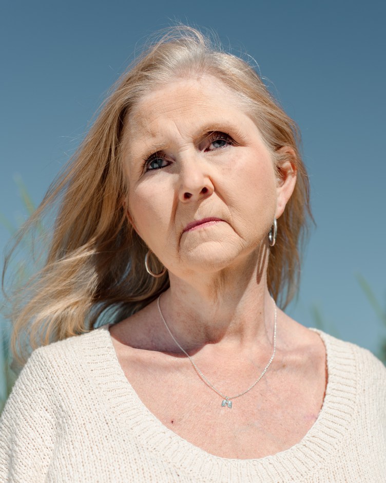 A woman with long blonde hair blowing in the wind faces the camera and stares off into the distance. She wears a white knit sweater, a silver necklace and silver hoop earrings. Out-of-focus plants appear behind her against a blue sky.