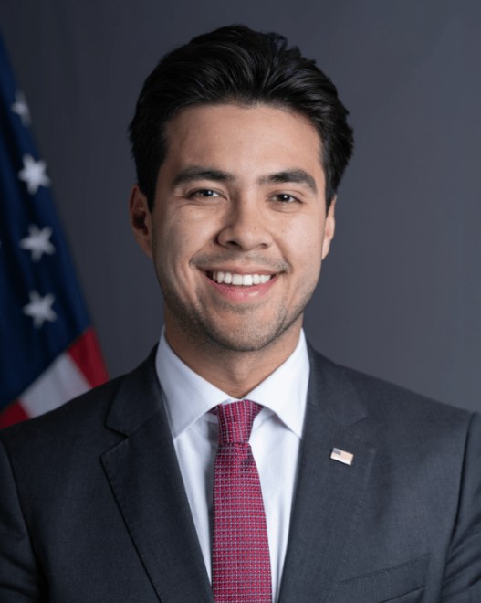 A man in a suit and tie smiles for his portrait. In the background there is a sliver of the American flag.