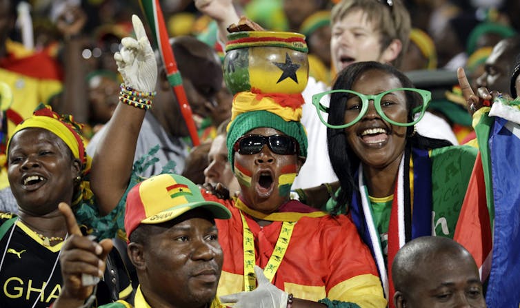 fans at a stadium cheering wearing clothing with the colours of the ghana flag. One is balancing a pot of their head.