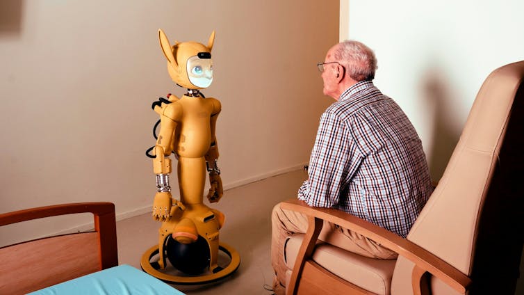 A yellow robot with a human-fox face stares at an older man who sits in a chair.