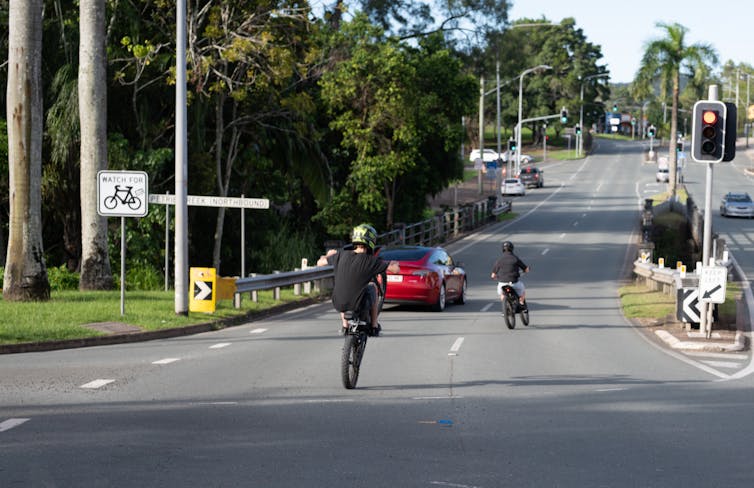 two e-bike riders, one doing a wheelie on a public road.