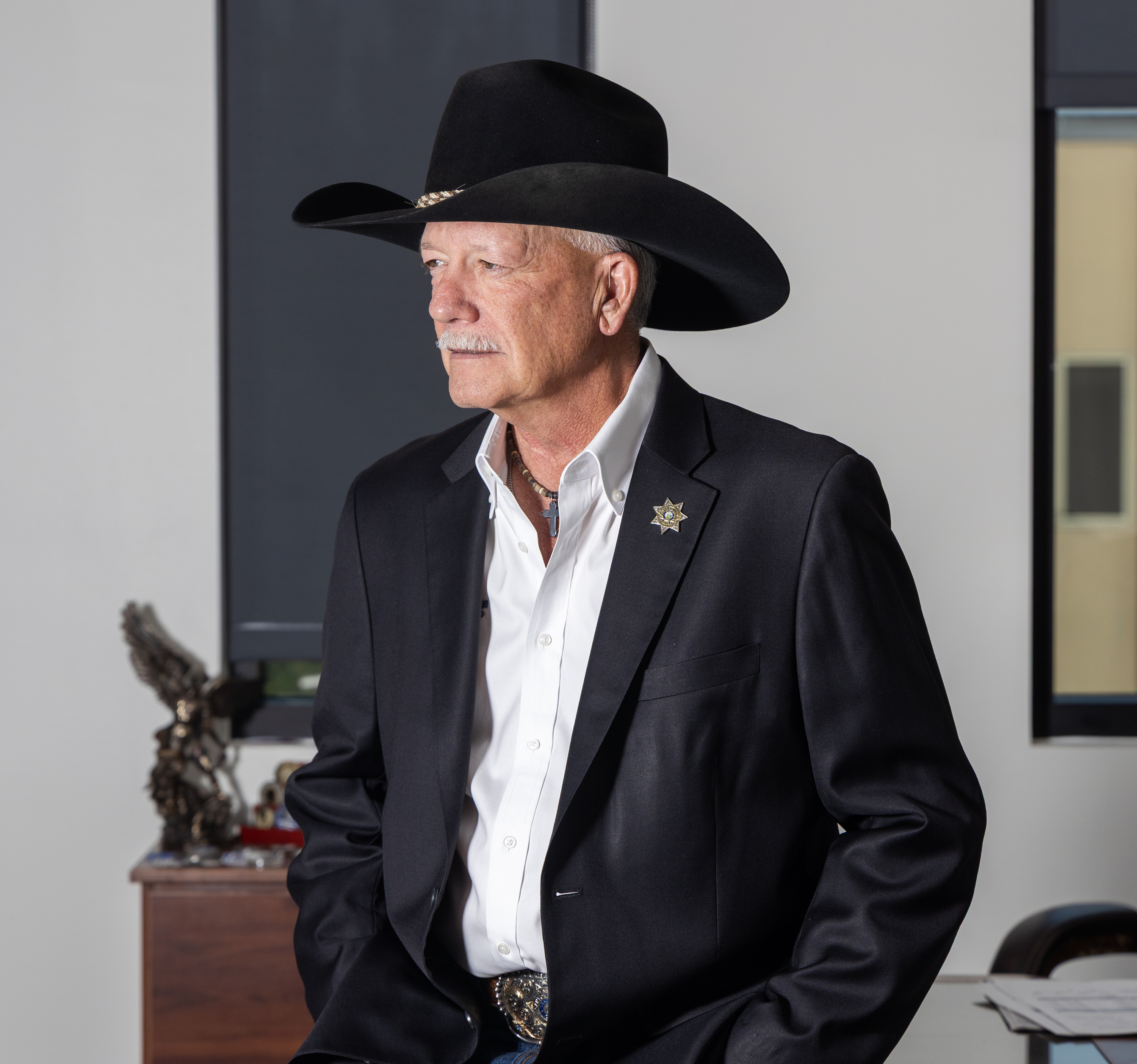 A man looks off frame toward his right in a room with vertical windows. He wears a black cowboy hat, a sheriff’s star pinned in the lapel of a black blazer, a large silver belt buckle and a silver cross on a necklace.