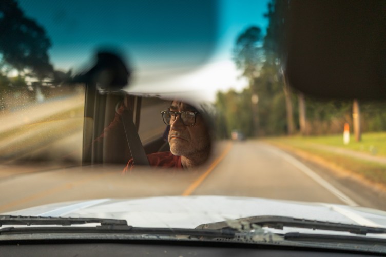 A man wearing glasses is reflected in the rearview mirror of the car he is driving.