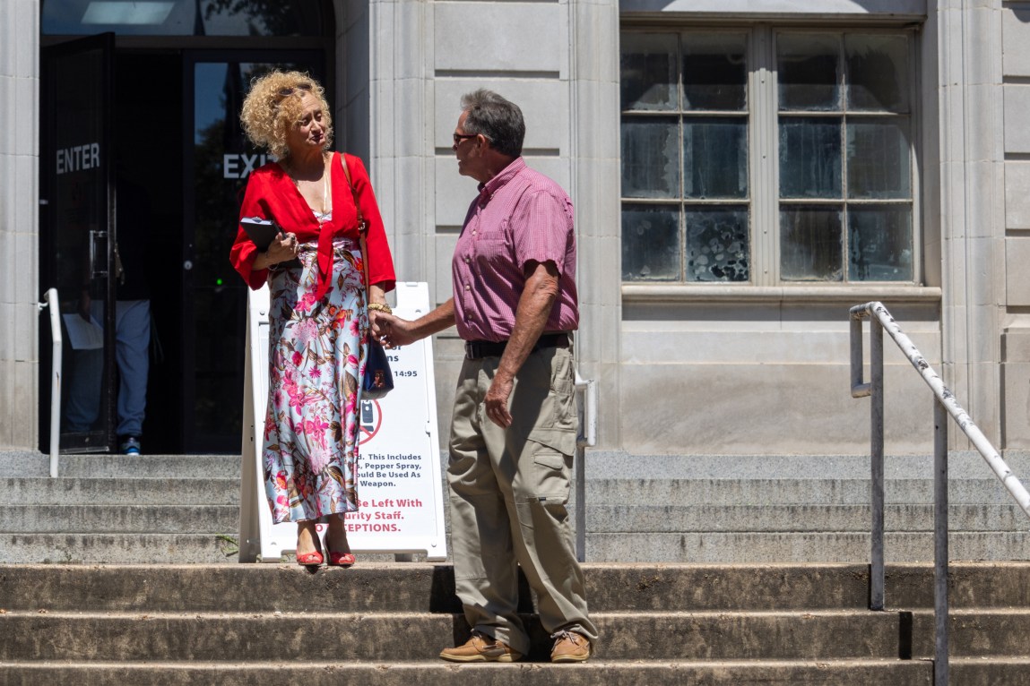 A woman stands on the steps of a courthouse building, holding back tears, while a man reaches out and holds her hand.
