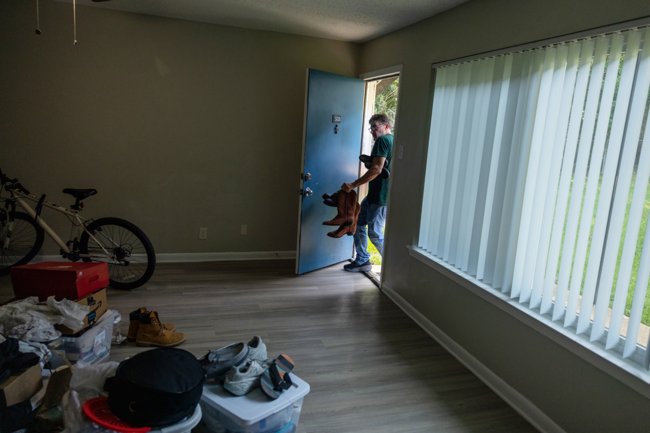 A man walks into the doorway of a simple apartment, carrying cowboy boots and other belongings. Boxes, a bicycle and other items are on the floor.