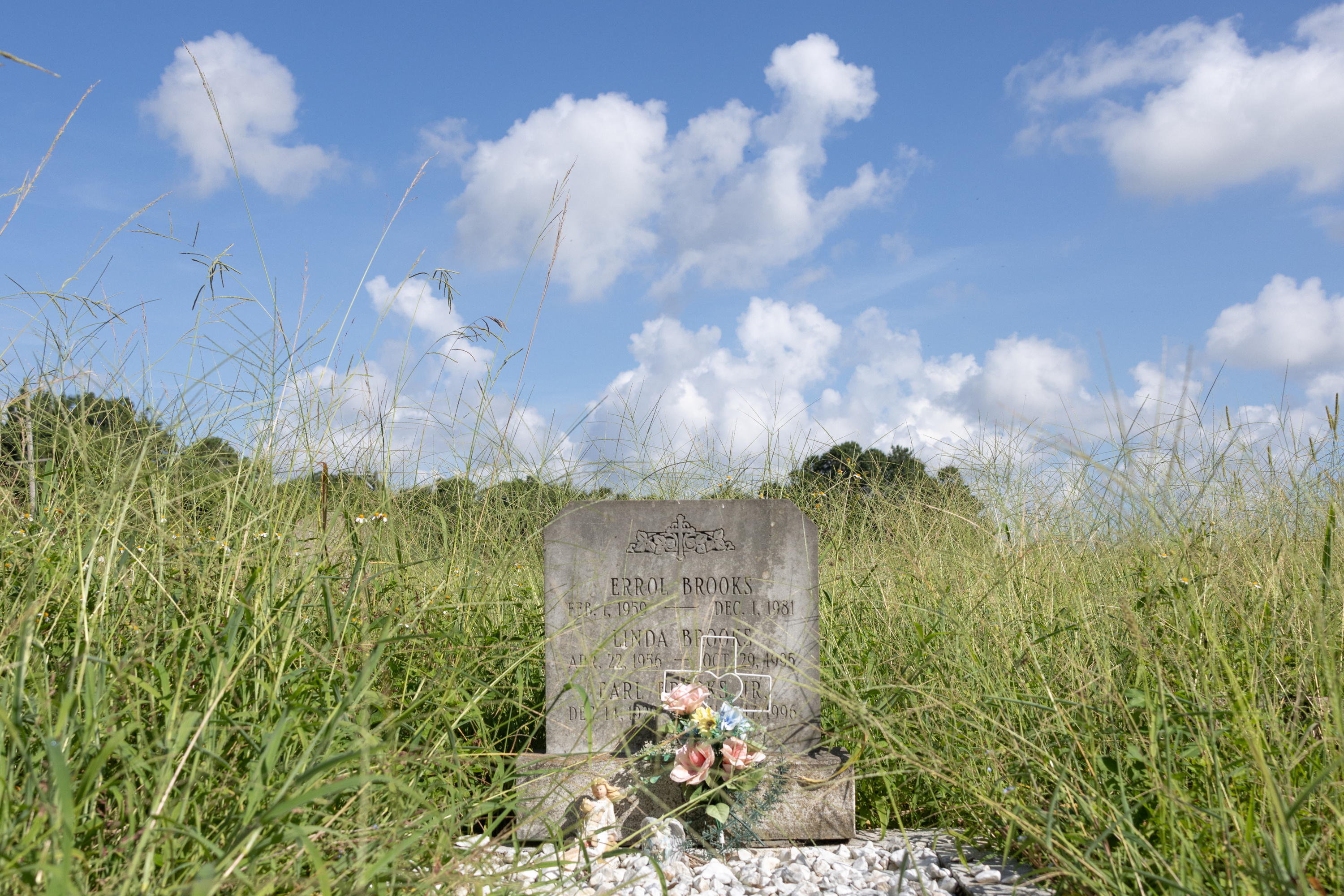 A gravestone with a cross etched on top and the words “Errol Brooks, Feb. 1 1959 — Dec. 1, 1981.” “Linda Brooks, Apr. 22, 1956 — Oct. 29, 1995,” and “Earl.” A bouquet of flowers and a cross obscure the last name. Two small angel statues also lean against the grave. Gravel sits in front of the grave, and long green grass grows behind the grave. The sky is blue with some clouds.