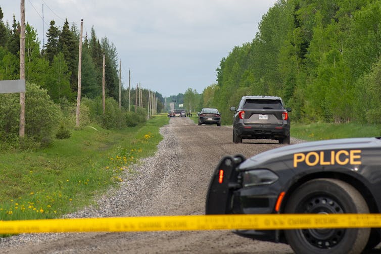 Three police cruisers on a rural road behind yellow police tape.