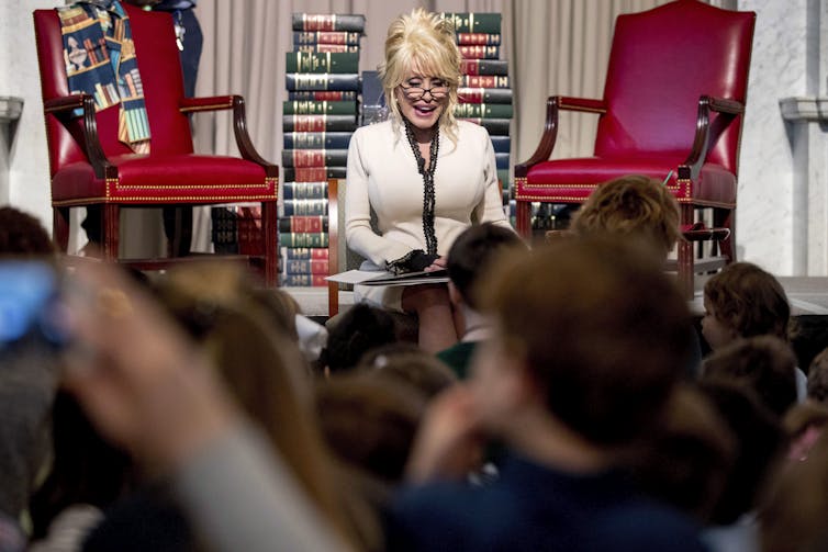 A middle-aged white woman with voluminous blonde hair reads a book to a crowd of seated children