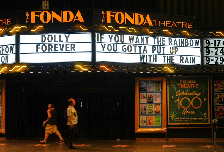 Pedestrians pass under marquee signs that say 'Dolly forever' and 'If you want the rainbow you gotta put up with the rain.'