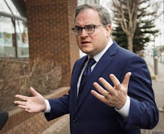 A man wearing glasses in a blue suit gestures as he speaks.