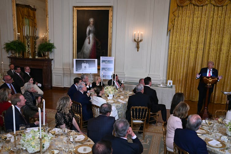 Donald Trump speaks to people in a ballroom with gold-framed paintings.
