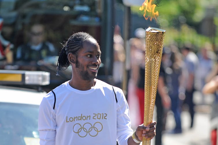 a young smiling man holds a lit gold torch. His t-shirt has the olympic logo and London 2012