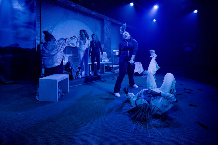 Five performers in various positions on a blue-lit stage, including one lying down on the floor and one dancing.