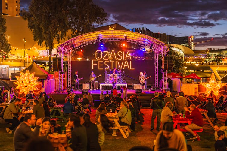 Front view of a stage at an arts festival, with tables full of people eating and chatting in front.