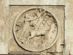 A round carving depicting a man in a horse-drawn chariot.