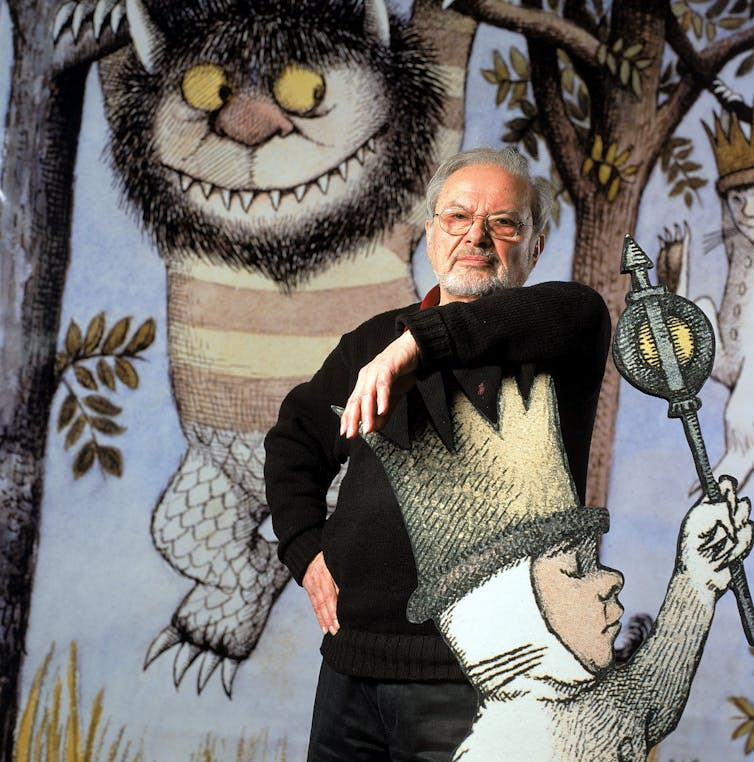A man stands and puts his arm against the crown of a large cutout cartoon child. Images of cartoon monsters hanging from trees are seen hanging behind him.