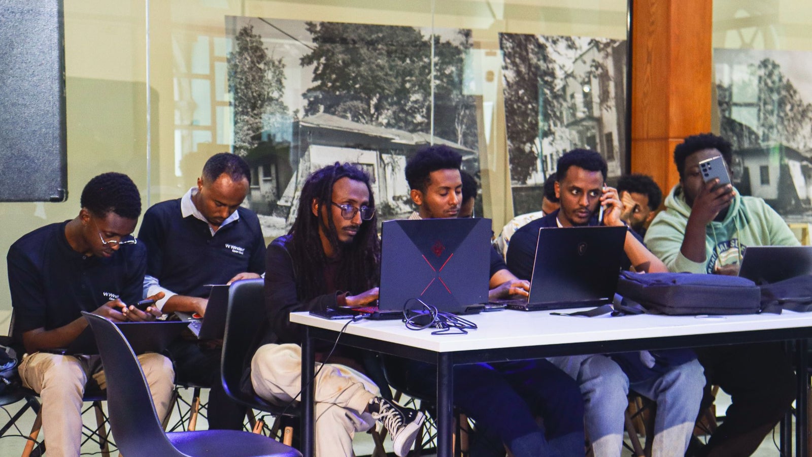 A group of young adults work at laptops and use phones in a modern indoor space, focused on their tasks