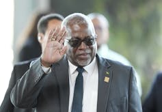 A Black man in a suit and sunglasses waves.