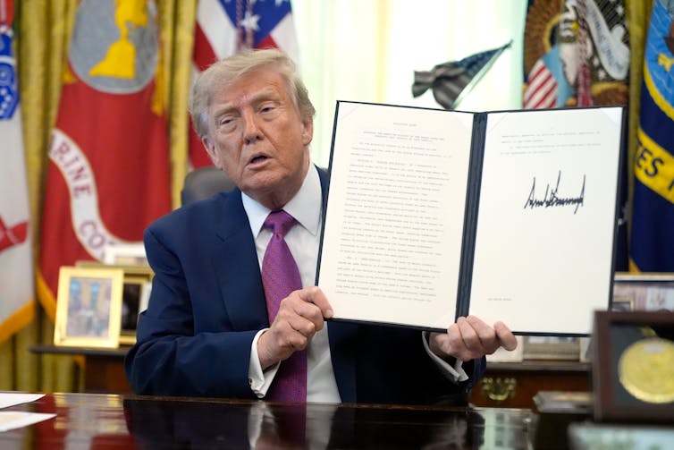 A man with yellow hair in a navy suit jacket and purple tie holds up a paper in a folder with a signature in it.
