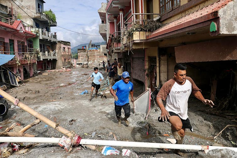 Three men running through a muddy street barefoot. Muddy vegetation is stuck to the second-floor balconies and mud can be seen most of the way up the side of a three-story building.